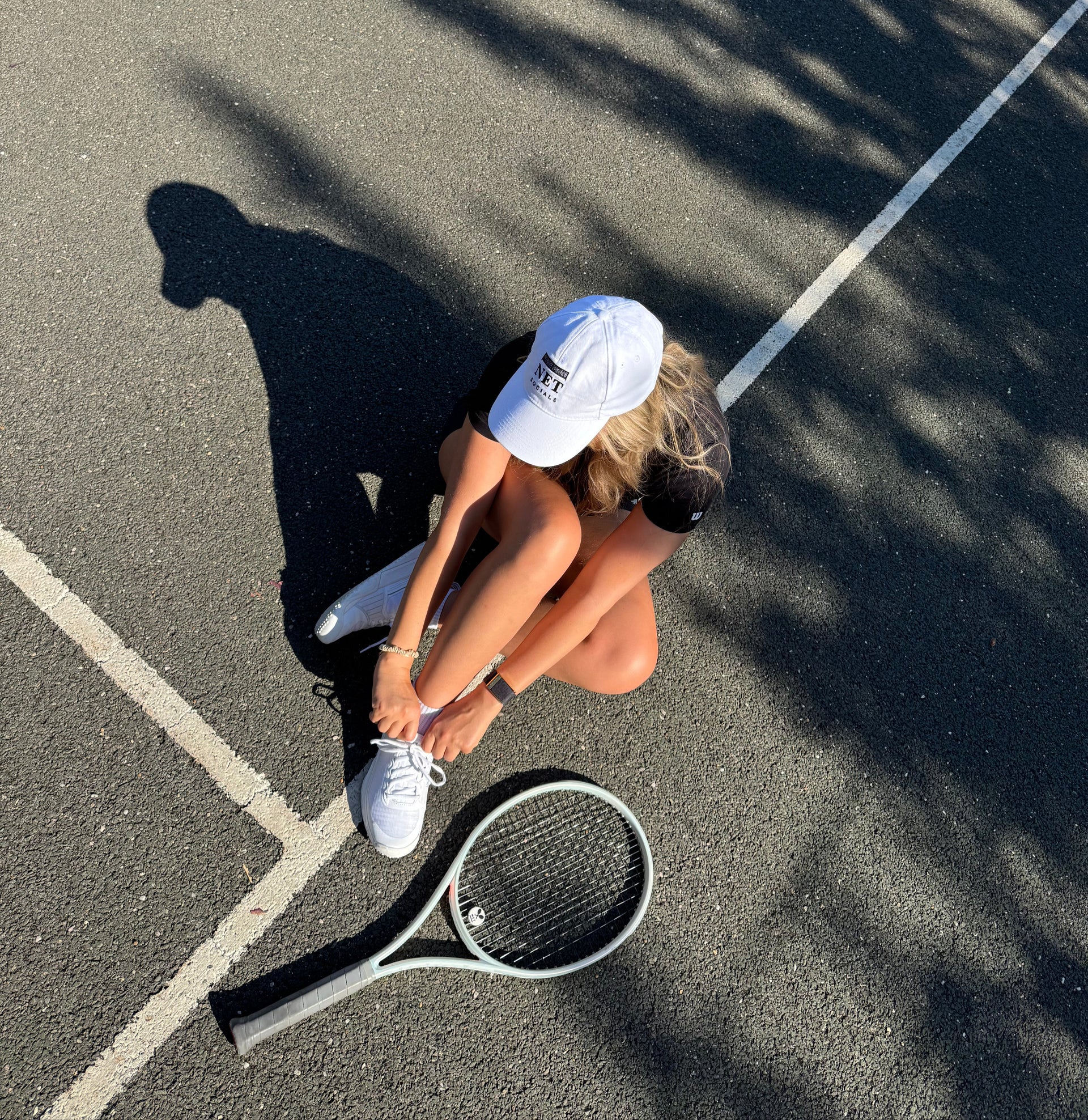 Person sitting on a tennis court with a racket, wearing a Net Collective x Net Socials cap and sunglasses.